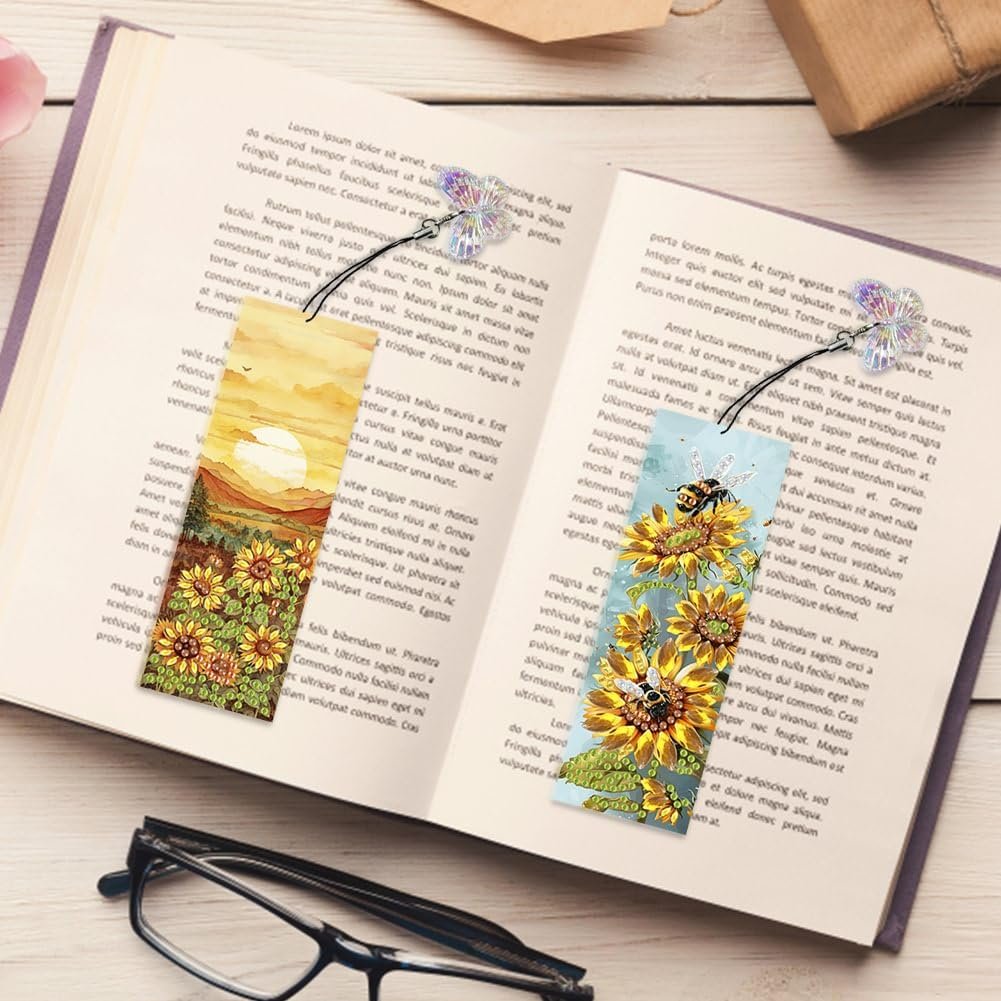 Two diamond painting bookmarks with sunflower and bee designs, placed in an open book with glasses nearby.