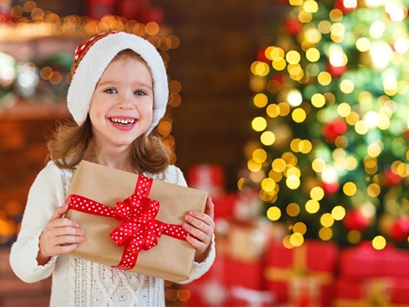 Smiling young girl in Santa hat holding a wrapped gift in front of a decorated Christmas tree