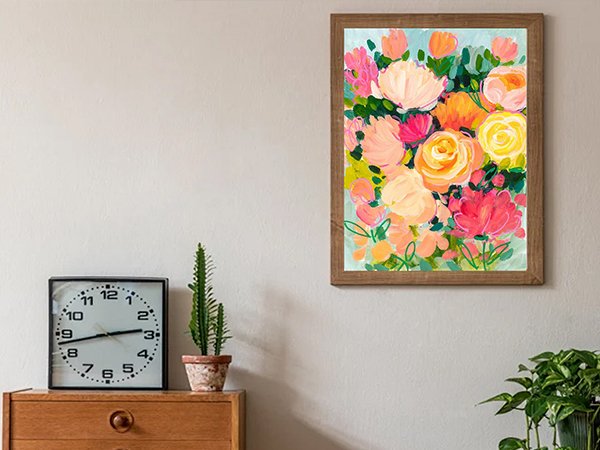 Framed abstract floral painting with pink, orange, and yellow roses hanging on a wall above a wooden dresser with a clock and plants.