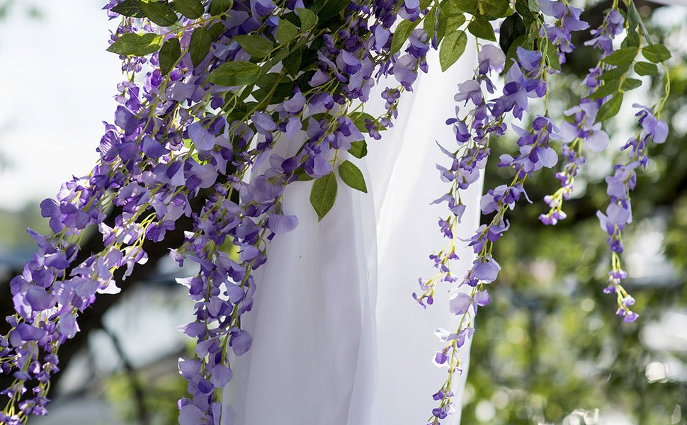 Purple artificial wisteria garlands with green leaves draped over white fabric, outdoors.