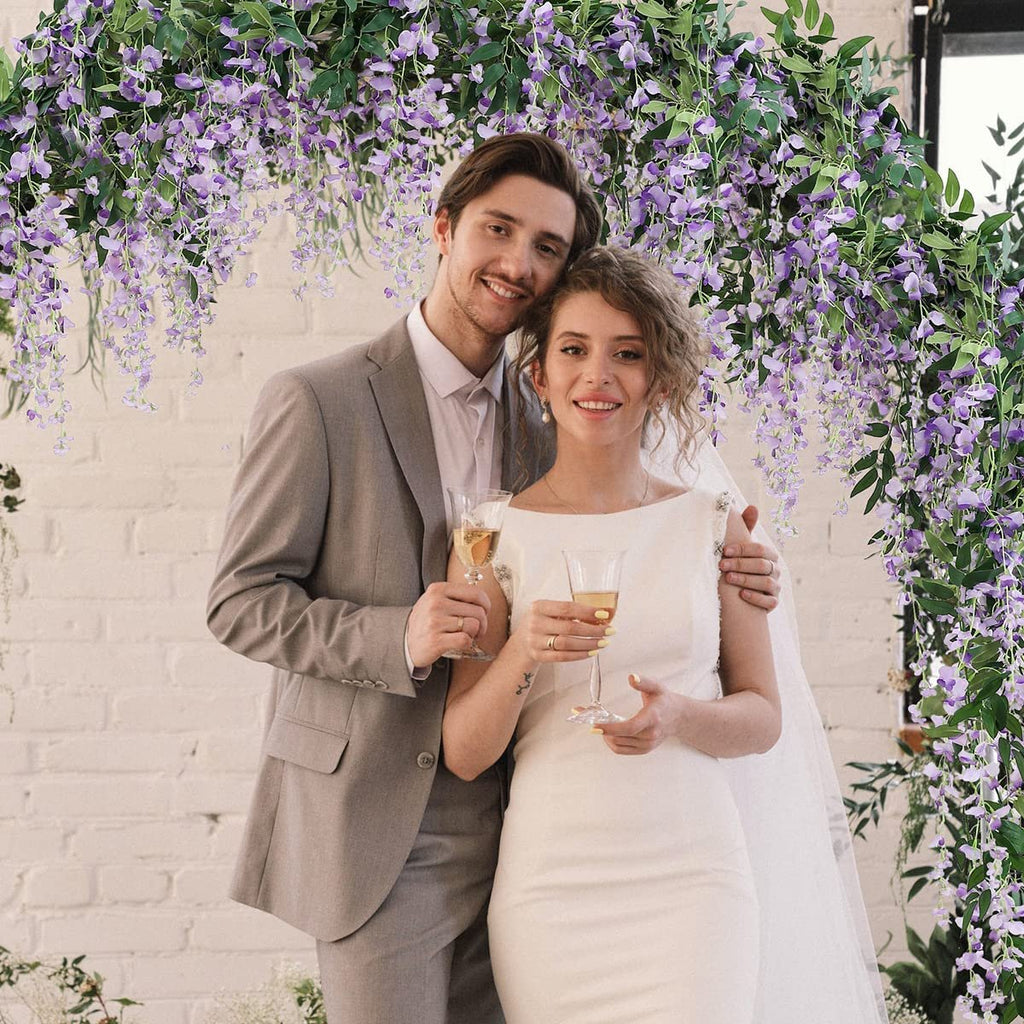 Couple celebrating under a purple wisteria flower archway, holding champagne glasses.