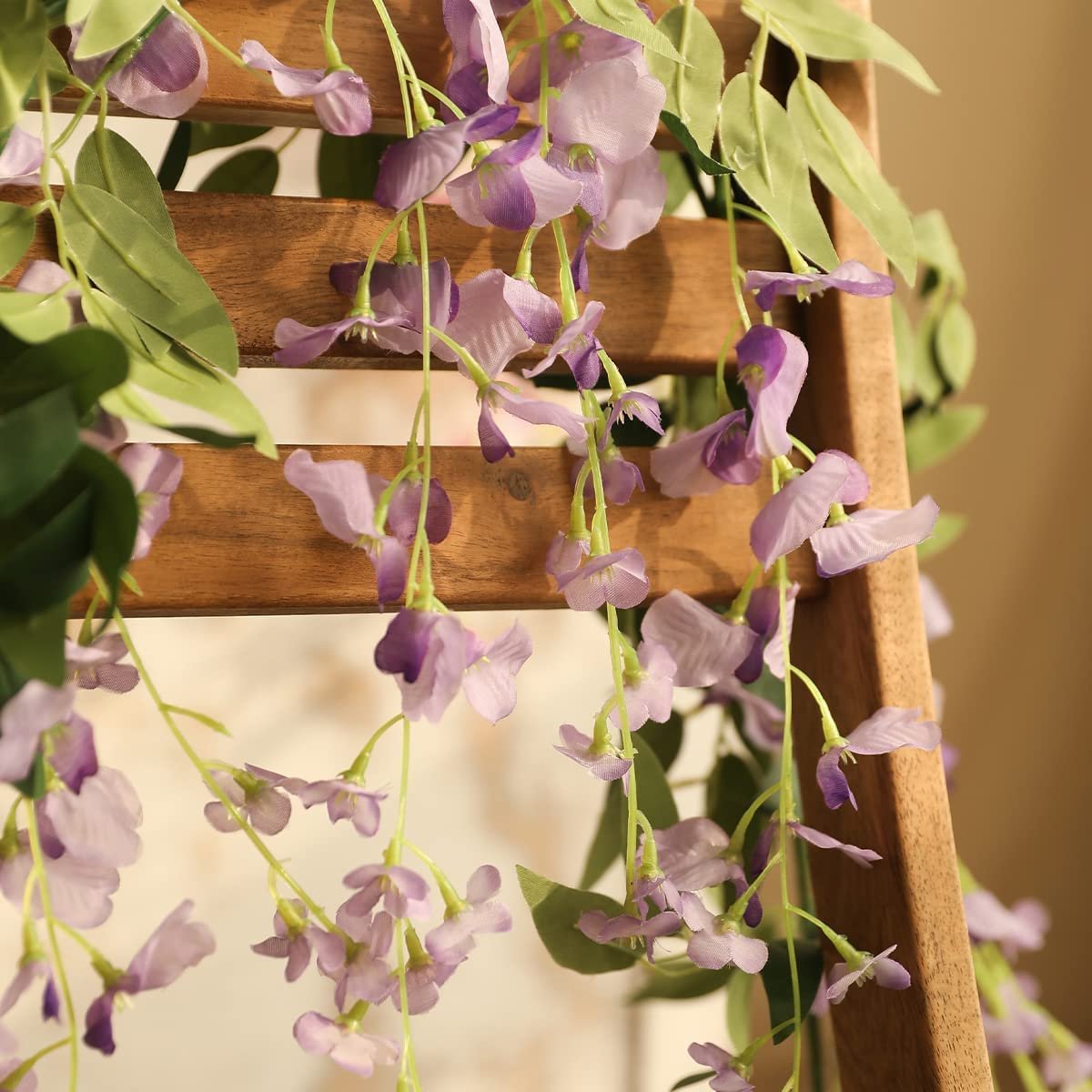 Purple artificial wisteria flower garlands with green leaves draped over a wooden ladder.