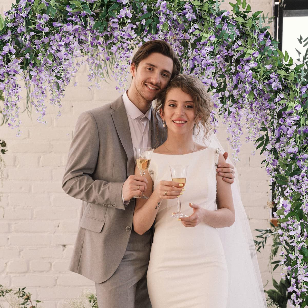Couple smiling under a purple wisteria flower archway, holding champagne glasses.