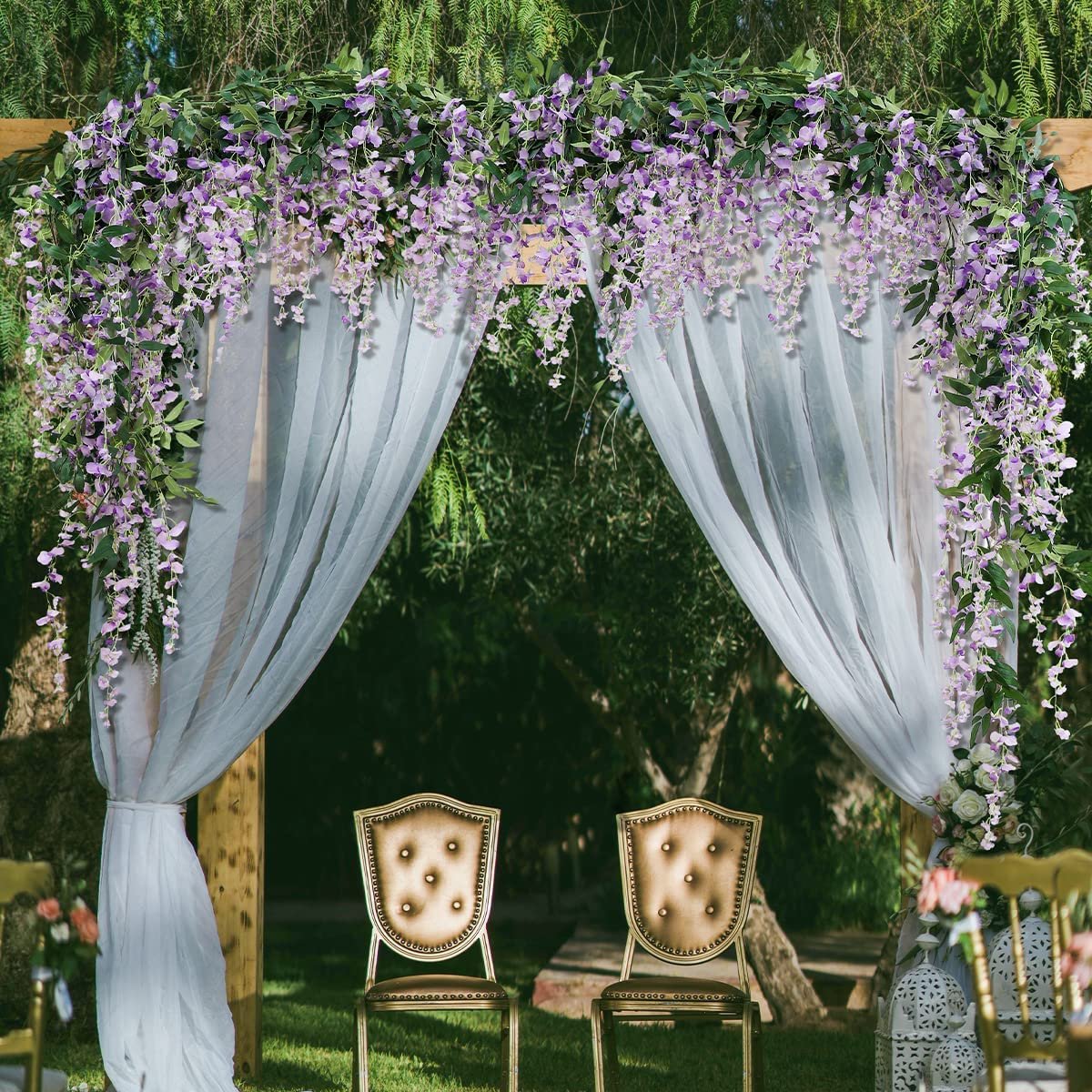 Wedding arch decorated with purple artificial wisteria garlands and white sheer curtains, with two ornate chairs underneath.