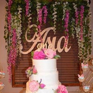 Three-tier white cake with pink flowers and butterflies, decorated with artificial wisteria garlands and a wooden 'Ana one' sign.