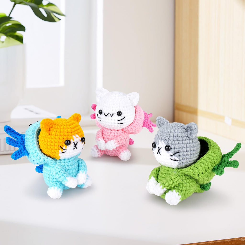 Three crocheted axolotl cat figures in blue, pink, and green outfits on a white surface with a plant in the background.