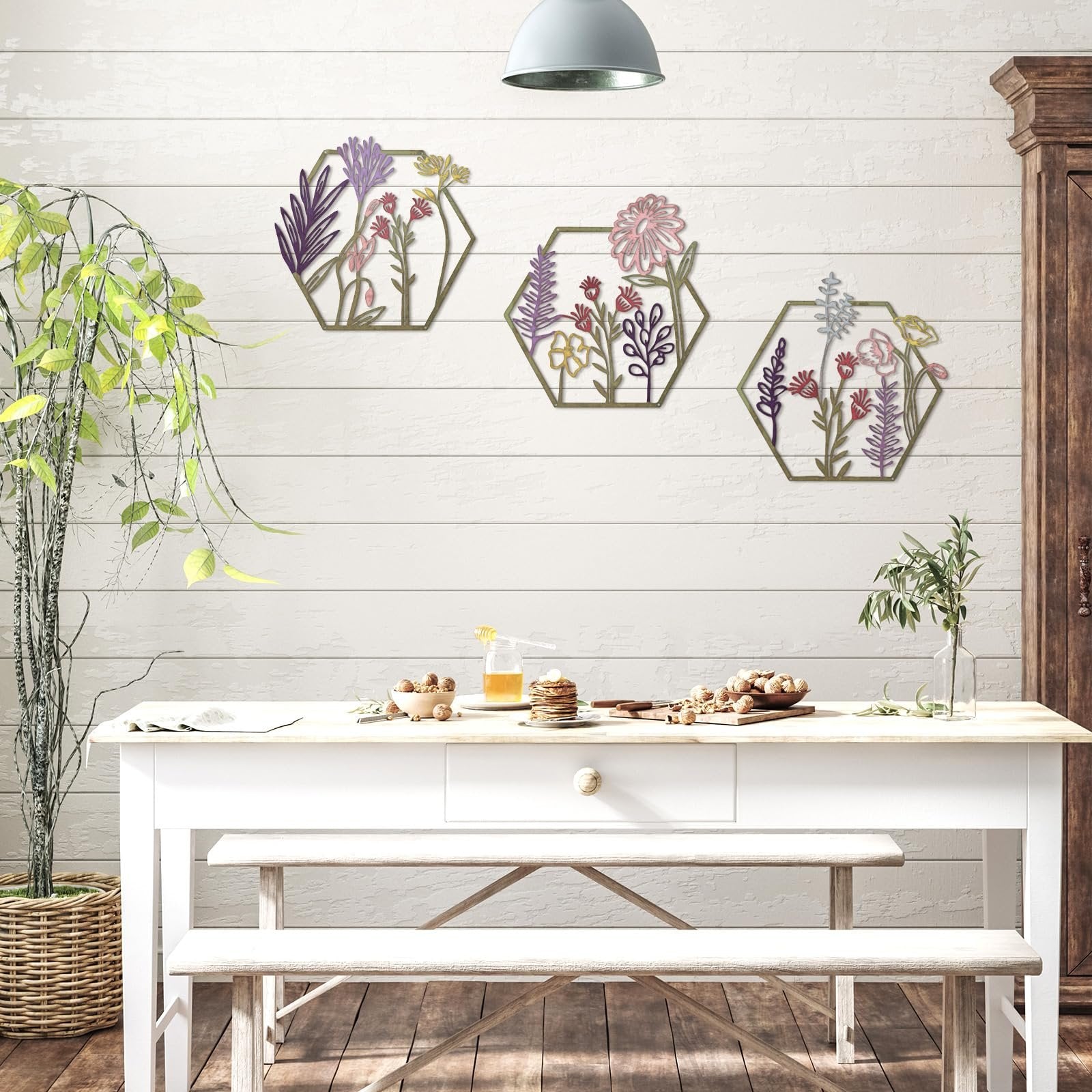 Three hexagonal metal flower wall decor pieces with colorful floral designs hanging on a white paneled wall above a table setting.