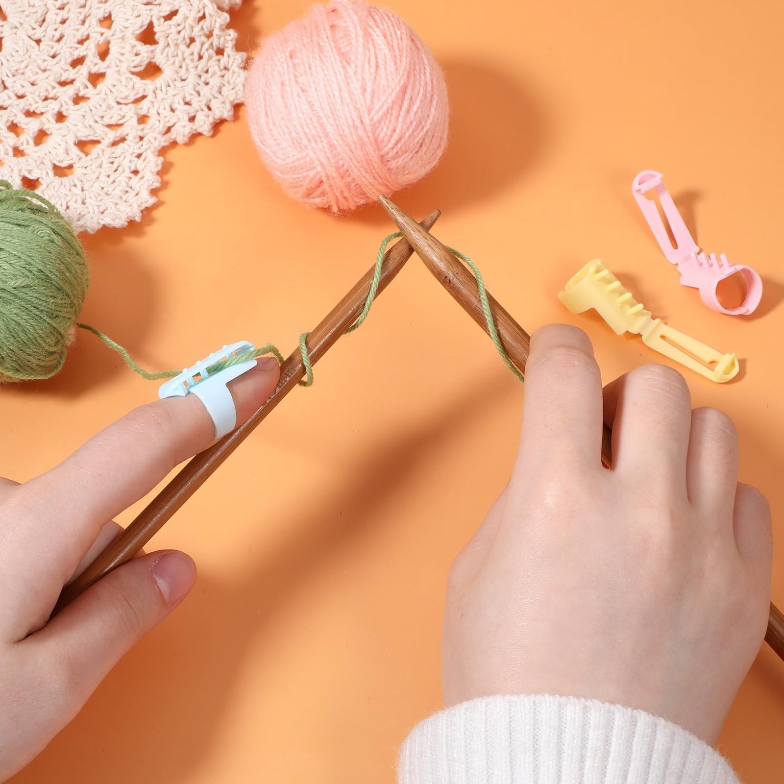 Hands knitting with yarn and finger tension tools, pink and green yarn balls, and a crocheted doily on orange background.