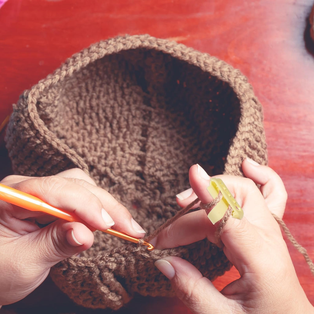 Hands crocheting a brown yarn project with an orange crochet hook and a yellow yarn guide ring.