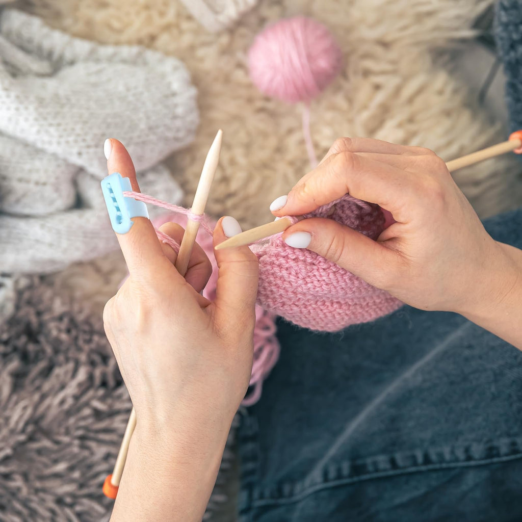 Hands knitting with pink yarn and a blue finger tension tool on a finger, surrounded by yarn balls and knitted fabric.