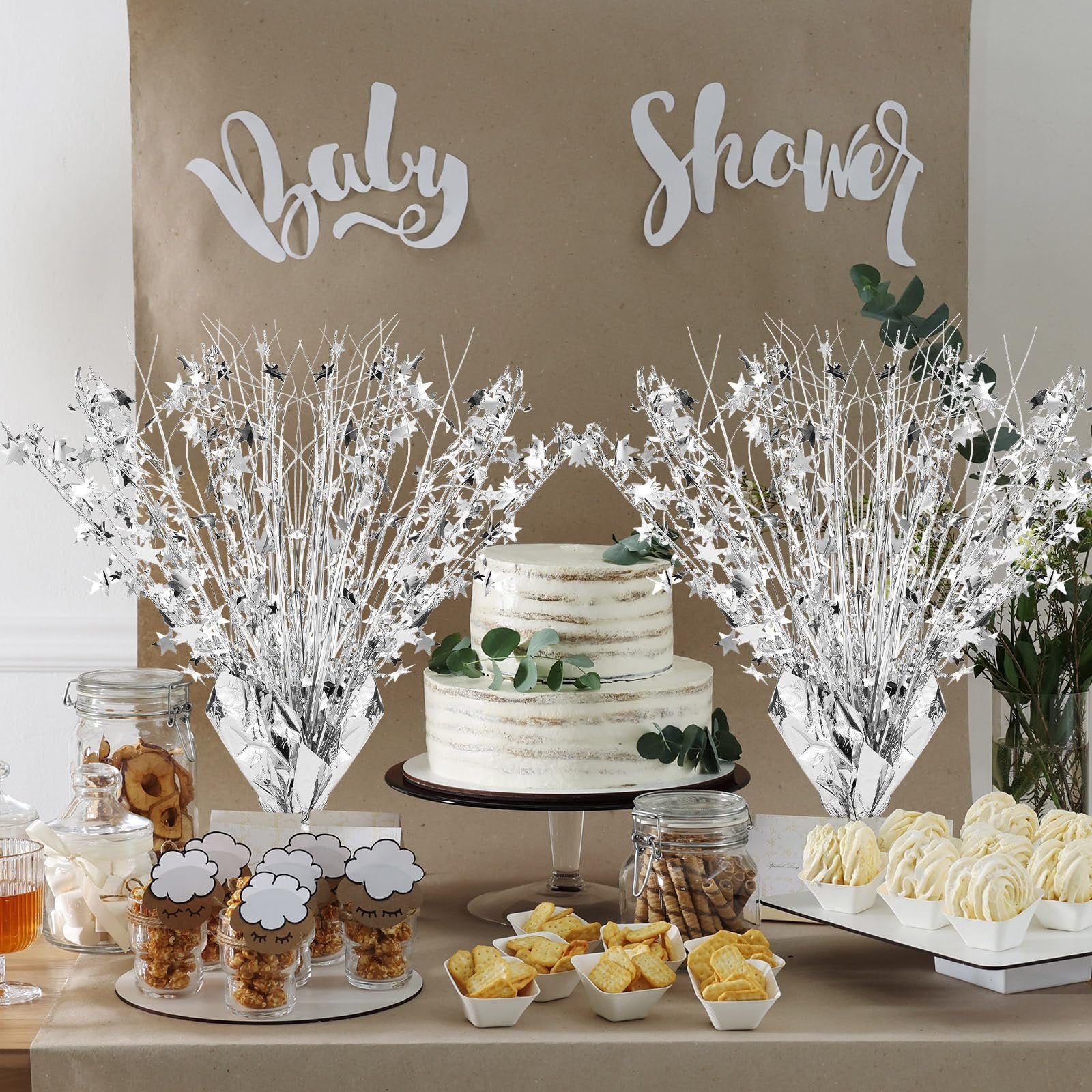 Two silver metallic star burst spray centerpieces flank a two-tier cake on a table with party snacks and decorations.