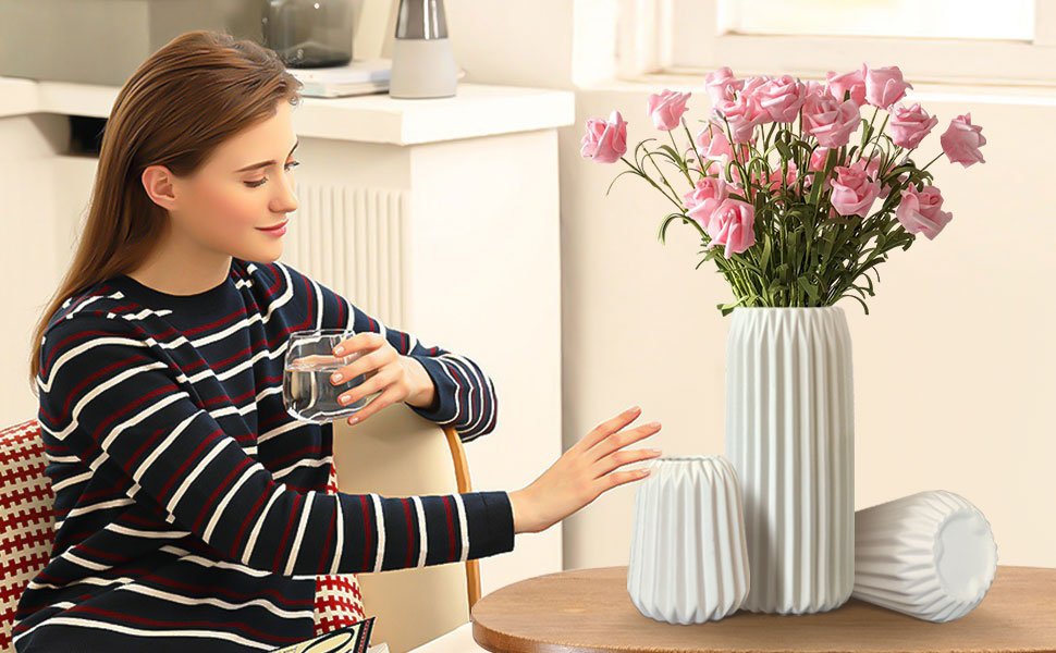 Woman holding a glass of water near a set of three white ribbed ceramic vases with pink flowers