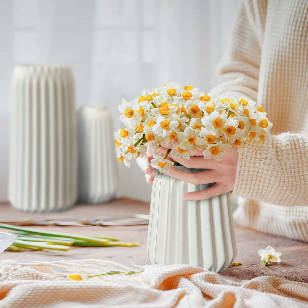 Hands arranging white daffodils in a fluted ceramic vase, with two other vases in the background.