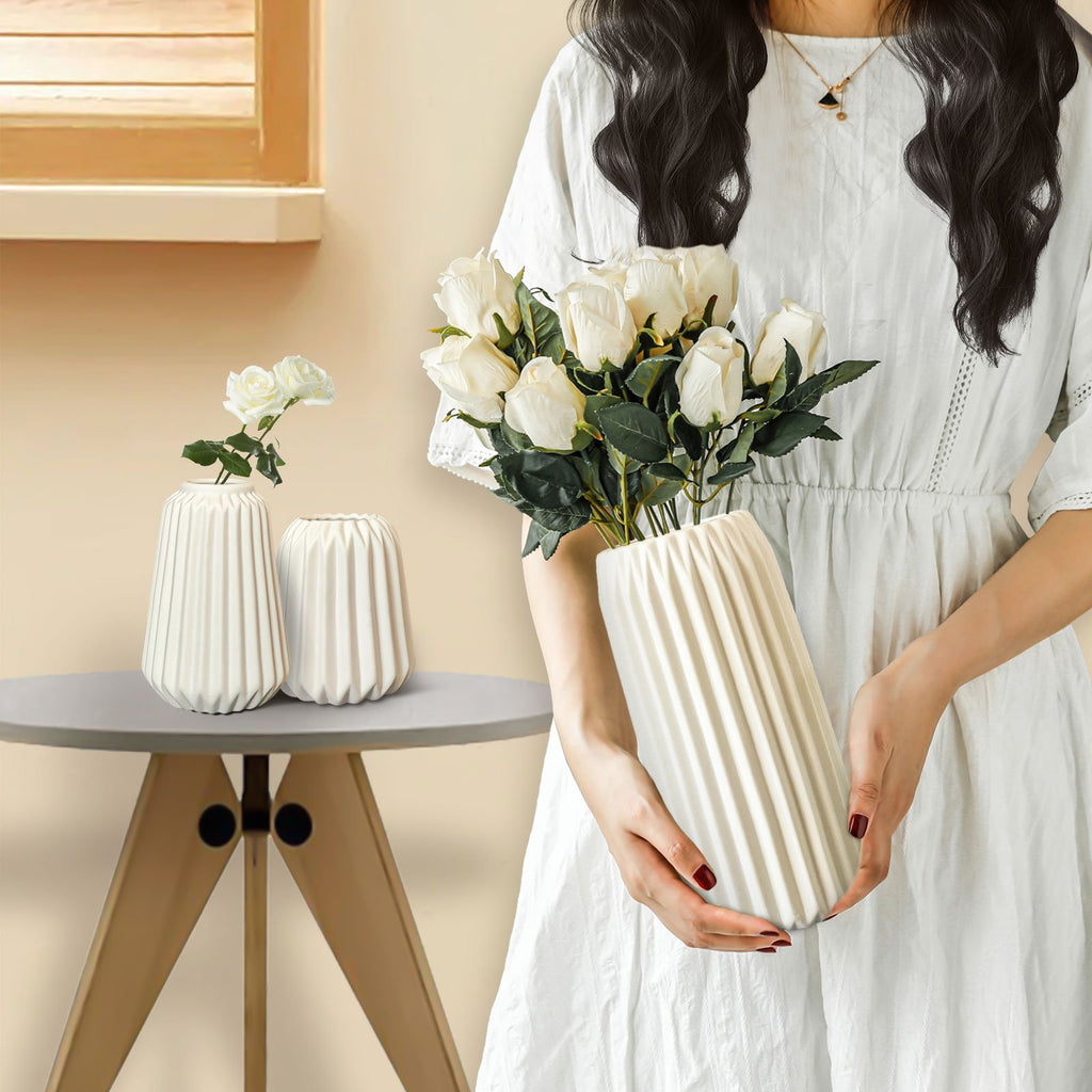 Woman holding a white pleated ceramic vase with white roses, two smaller vases on a table nearby.
