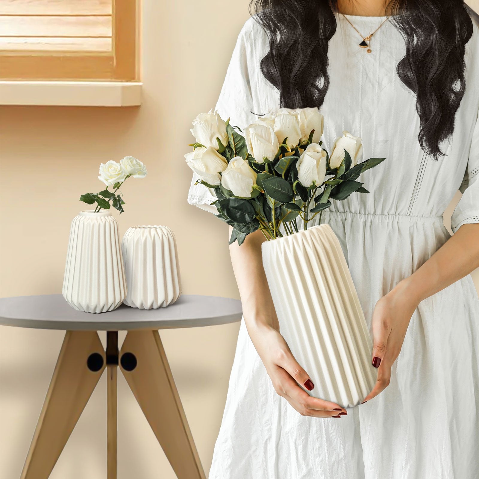 Woman holding a white pleated ceramic vase with white roses, two smaller vases on a table nearby.
