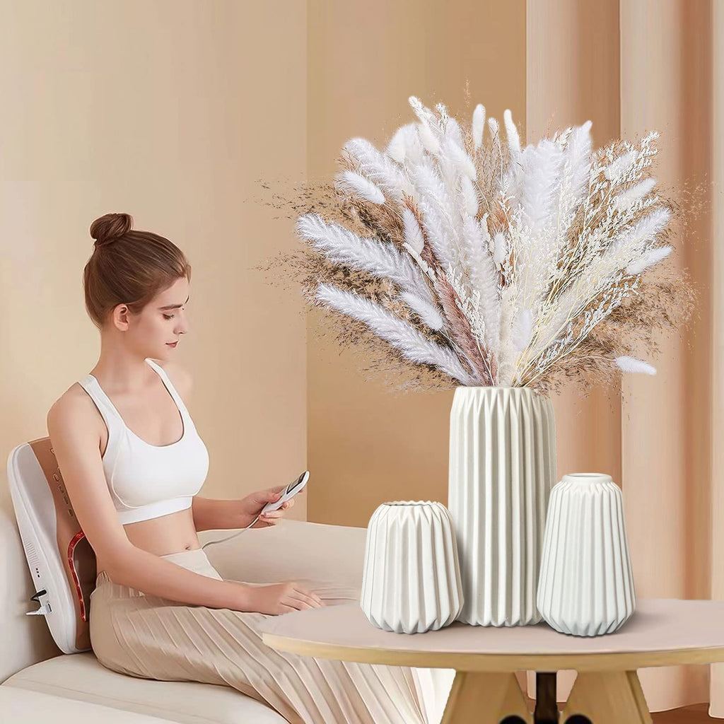 Woman sitting on a couch holding a remote, with a set of three white ceramic vases filled with dried flowers on a table in front of her.