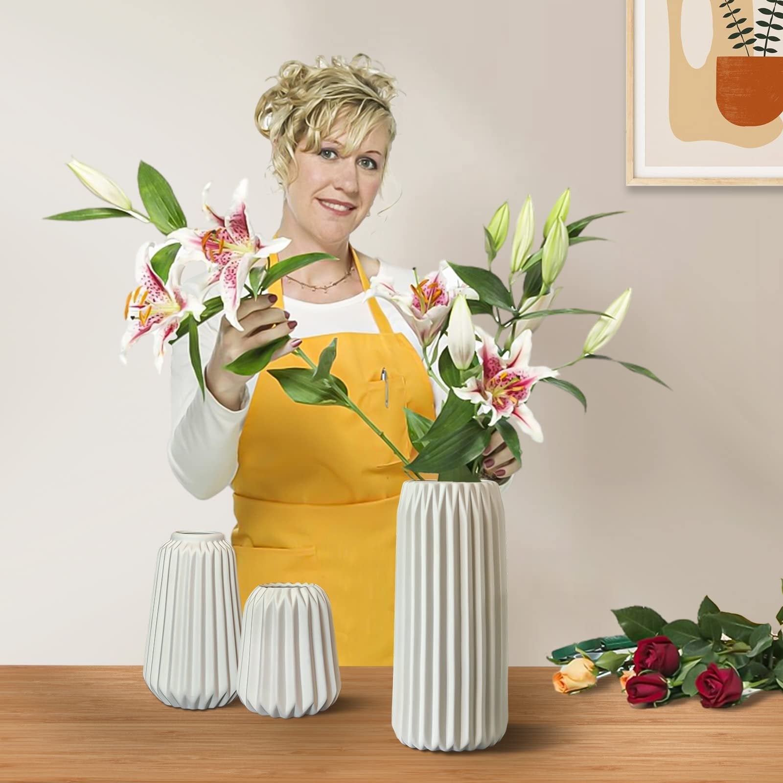 Woman in yellow apron arranging lilies in three white ribbed ceramic vases on a wooden table