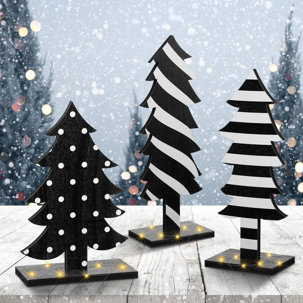 Three wooden Christmas trees with black and white patterns on a rustic wooden table with a snowy forest background and bokeh lights.