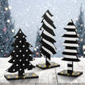 Three wooden Christmas trees with black and white patterns on a rustic wooden table with a snowy forest background and bokeh lights.