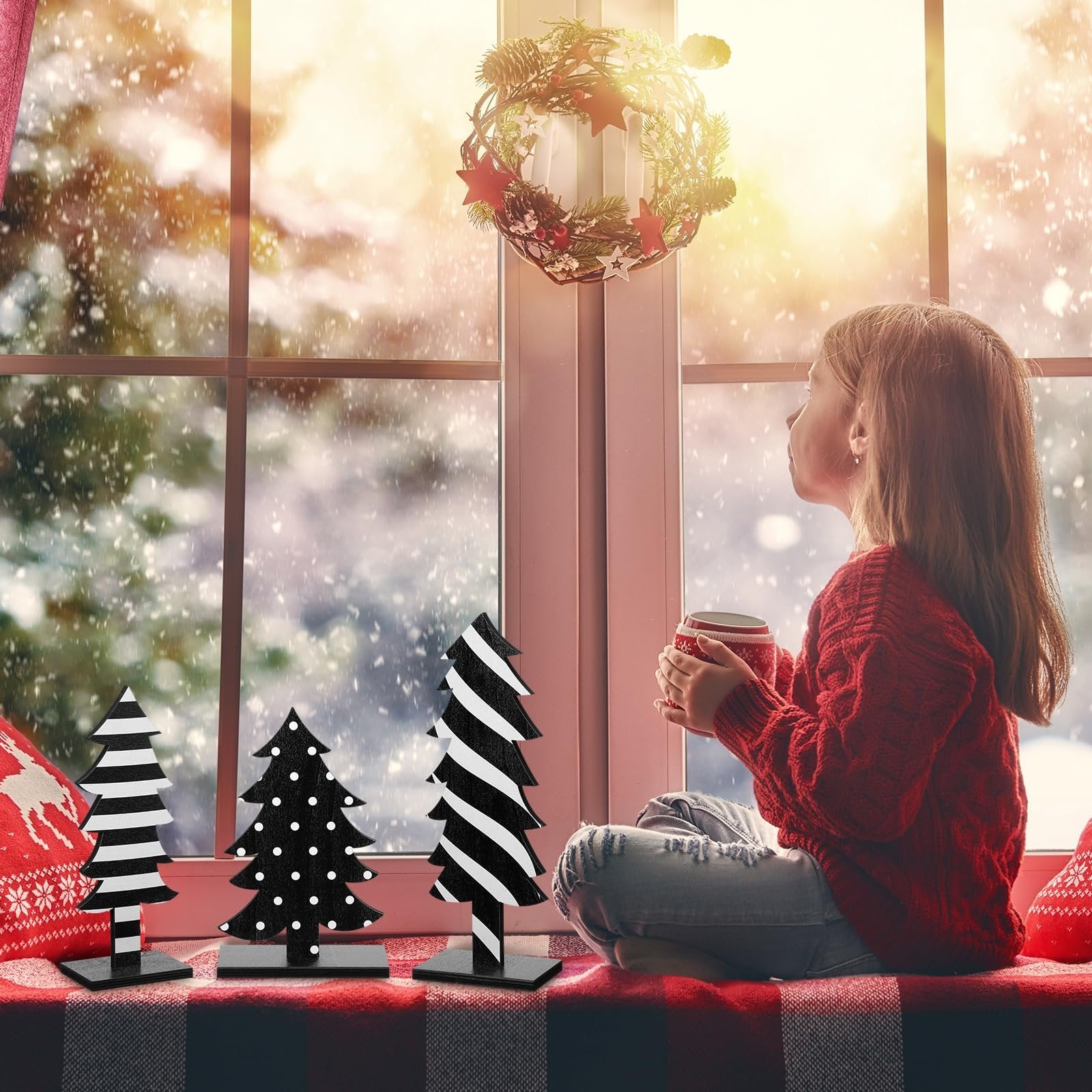 Girl in red sweater holding mug by window with wooden Christmas trees and wreath, snowy day