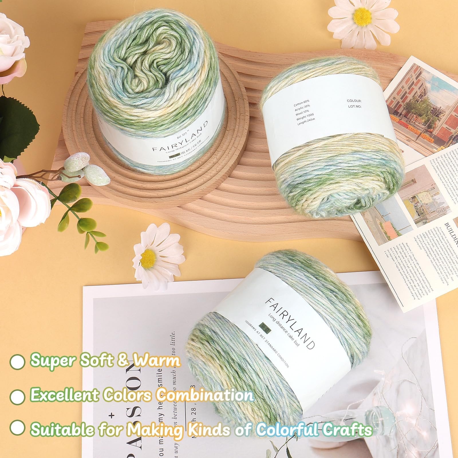 Three skeins of gradient cake yarn in green, blue, and yellow hues, arranged on a wooden tray with decorative flowers and printed materials.
