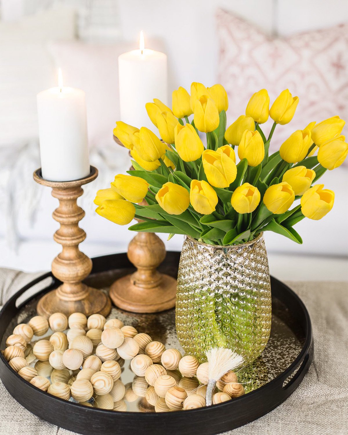Yellow artificial tulips in a textured vase on a tray with candles and wooden beads