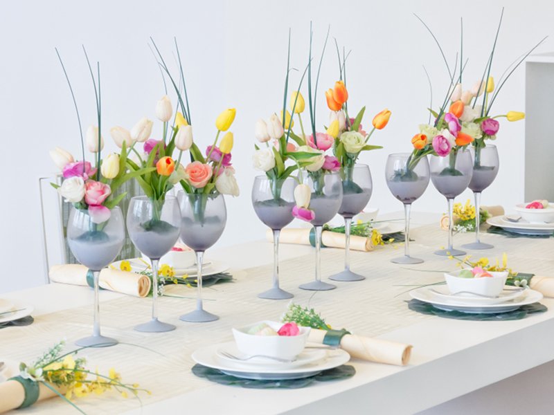 Table setting with multiple arrangements of artificial tulips in wine glasses, decorated for an event.