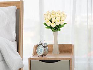 Bouquet of cream artificial tulips in a white vase on a wooden nightstand next to a bed.