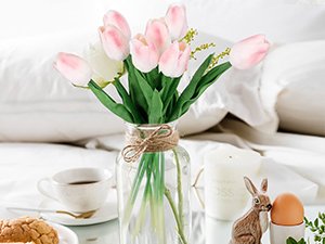 Bouquet of pink and white artificial tulips in a glass vase with twine, on a table with a coffee cup and Easter decor.