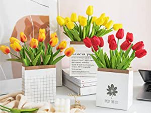 Three vases of artificial tulips in red and yellow, arranged on a desk with books and a laptop.