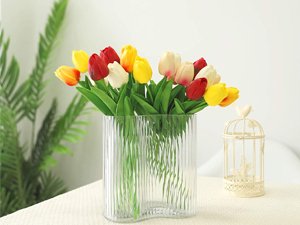 Bouquet of artificial tulips in red, yellow, and white in a clear ribbed vase on a table with greenery and a birdcage.