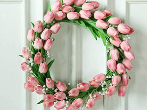 Pink artificial tulip wreath with green leaves and white filler flowers, hanging on a white paneled door.