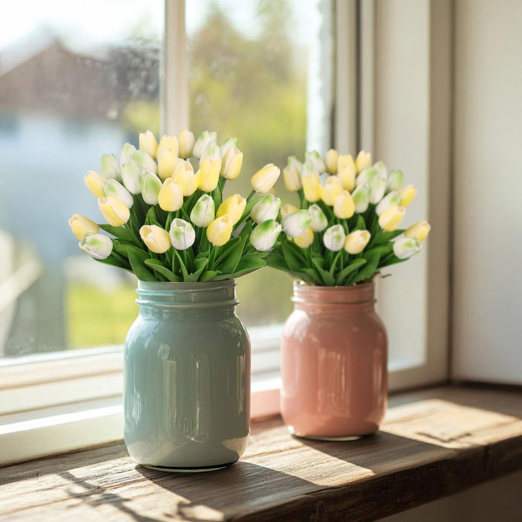 Two vases of artificial yellow and green tulips on a wooden windowsill with sunlight streaming through a window.