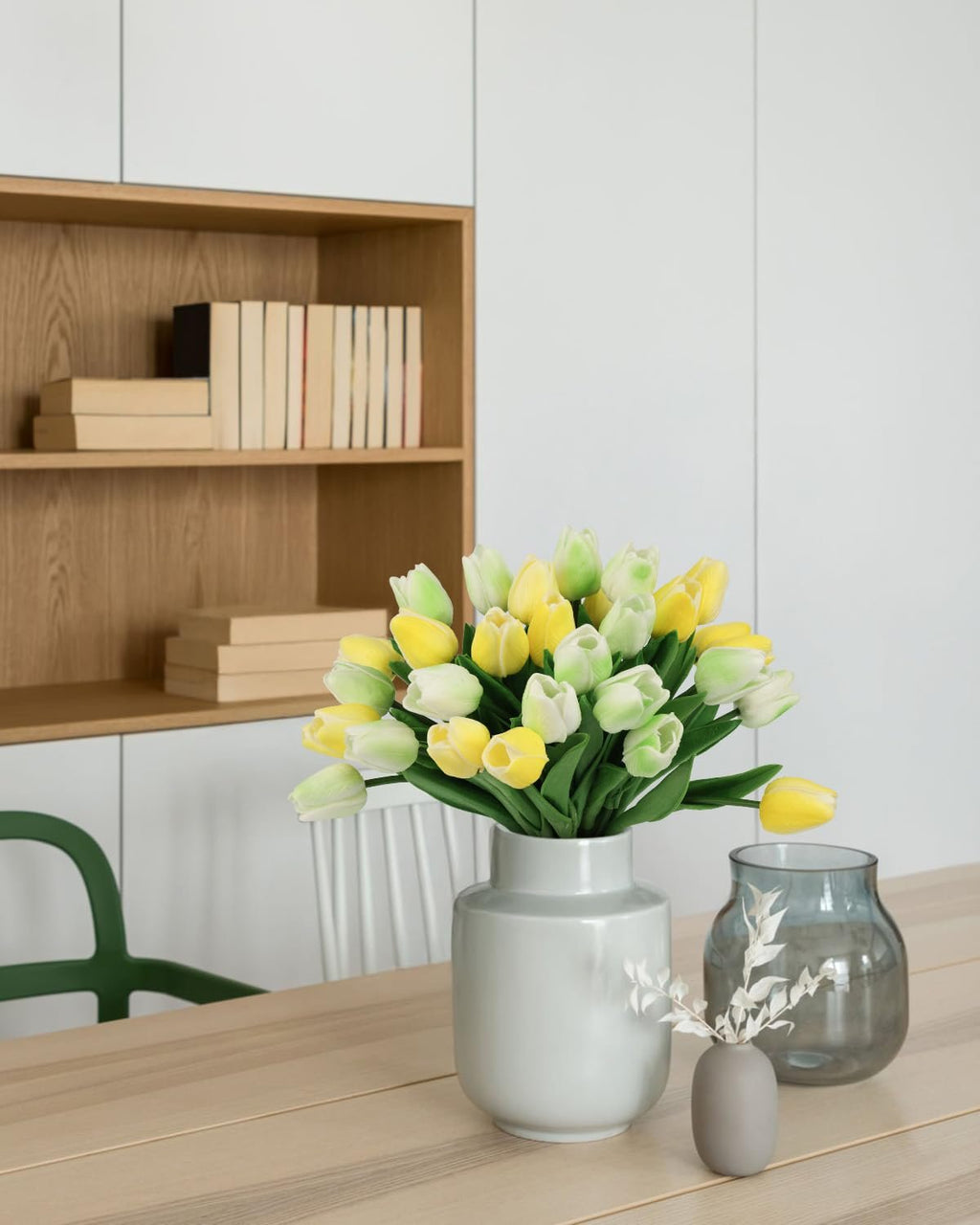 Bouquet of yellow and green artificial tulips in a vase on a wooden table, with books on a shelf in the background.