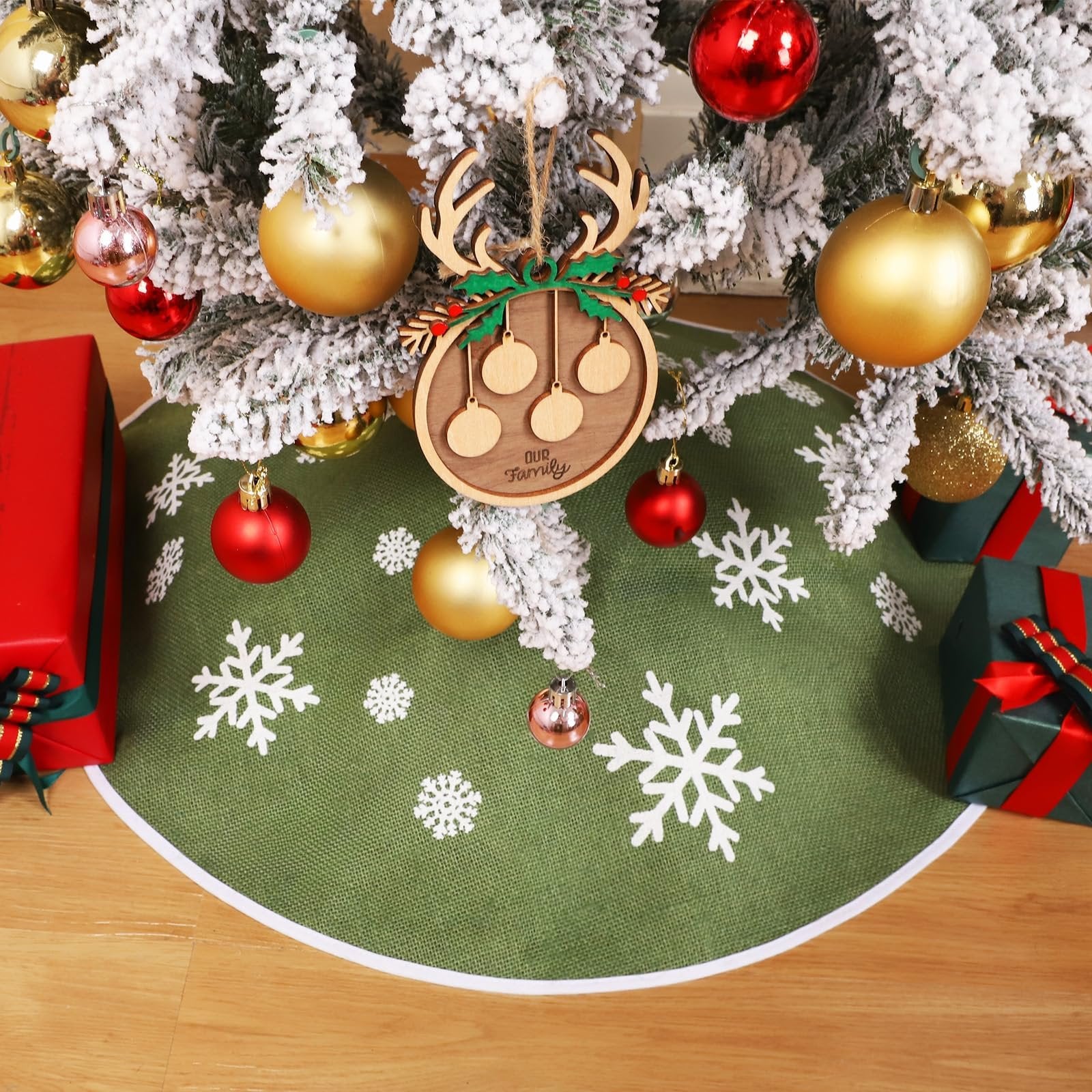Green burlap Christmas tree skirt with white snowflake print shown around the base of a decorated tree with gifts.