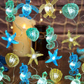 Nautical string lights with sea turtle, seahorse, starfish, and shell figurines on a wooden surface with a candle and netting.