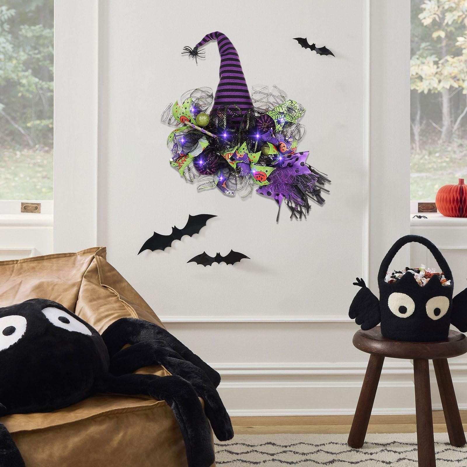 Halloween witch hat wreath with purple lights and mesh decor hanging on a wall, with a spider plush and candy bucket nearby.