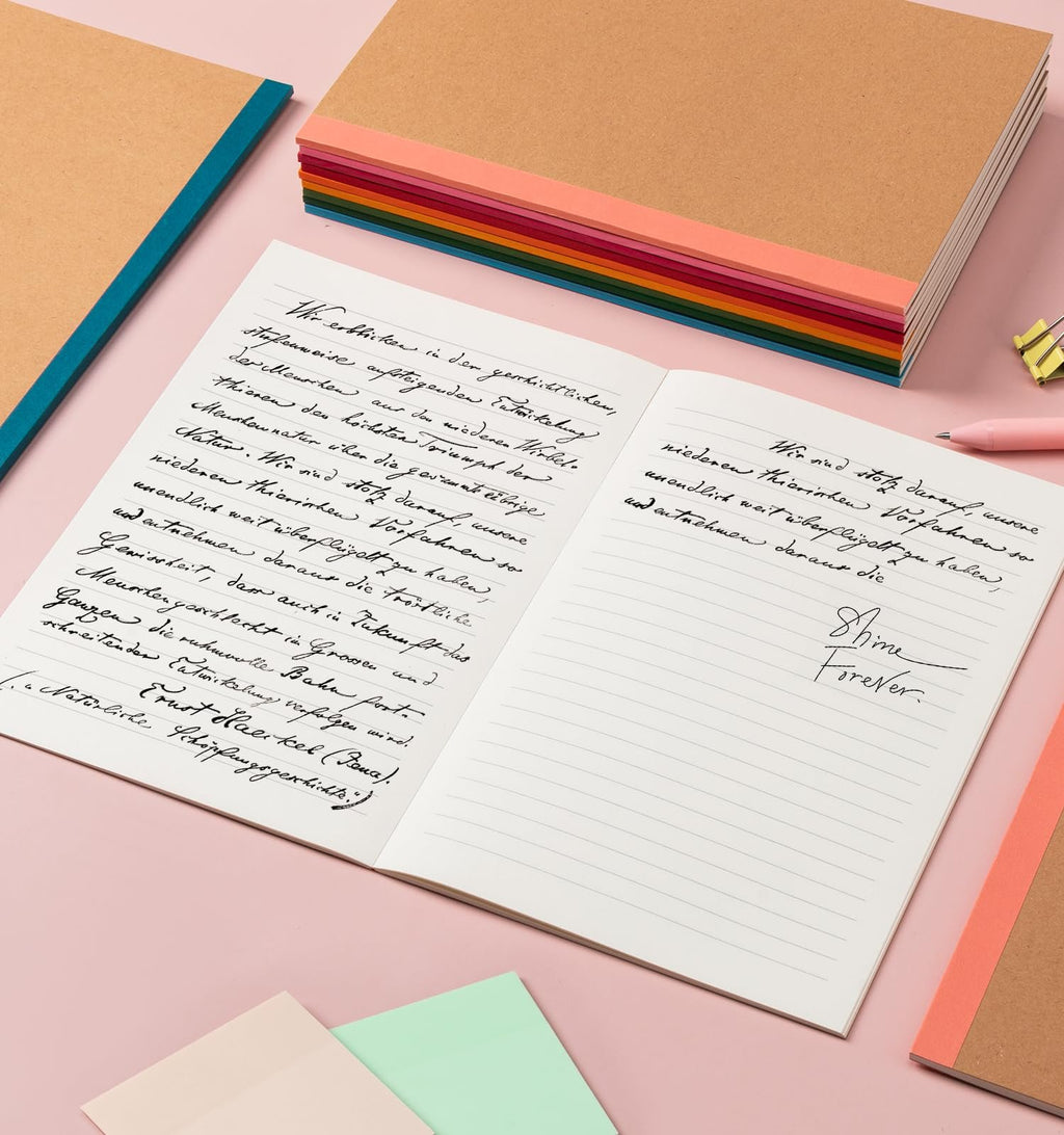 Open notebook with handwritten text and a stack of colorful A5 notebooks in the background.