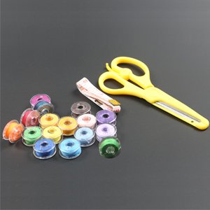 30-pack transparent plastic sewing machine bobbins with assorted thread colors and a yellow pair of scissors and tape measure on a dark gray surface.