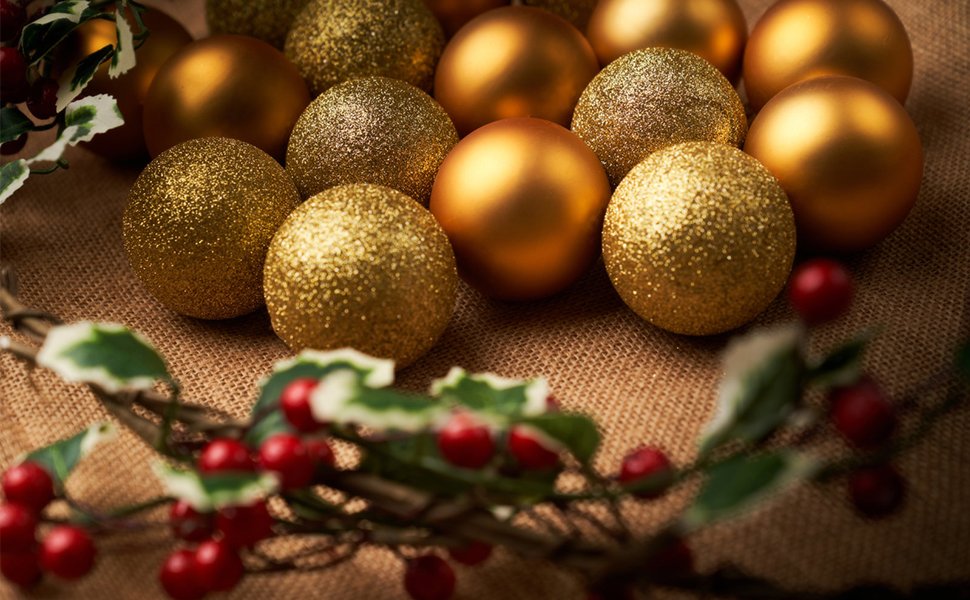 Gold and glitter Christmas ball ornaments arranged on burlap with a sprig of holly and berries in the foreground.