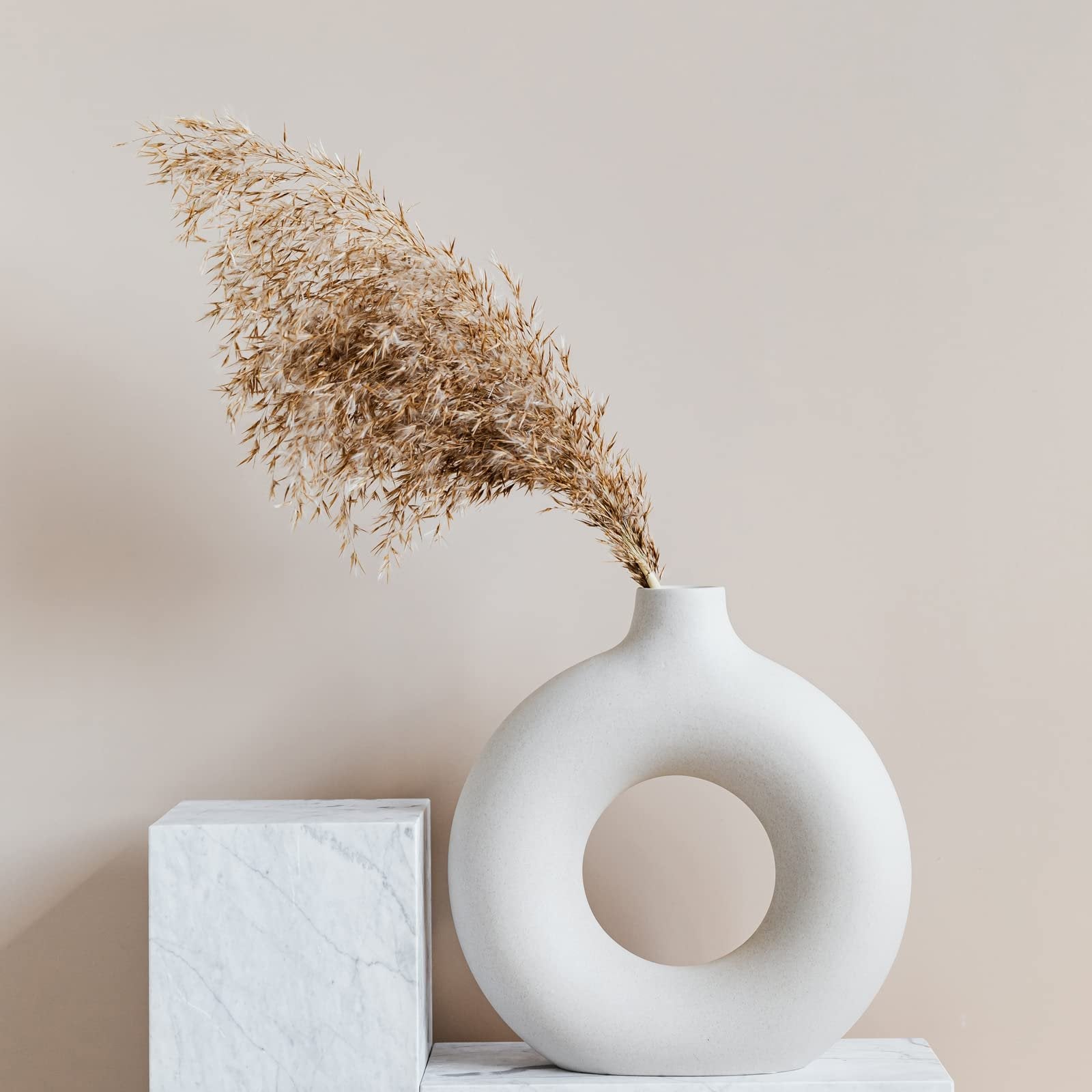 Dried pampas grass in a modern white donut-shaped vase on a marble pedestal.
