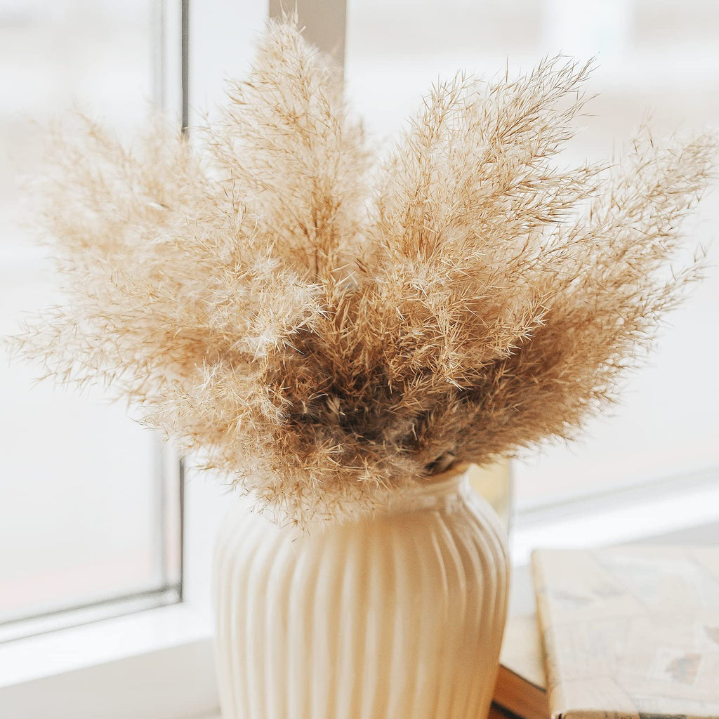 Close-up of a vase filled with dried pampas grass, placed on a windowsill next to a book.