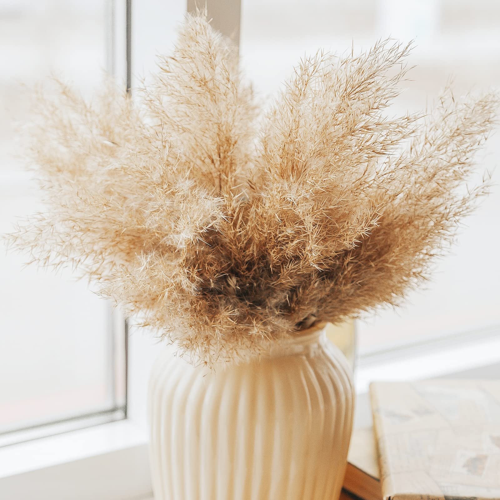 Close-up of a vase filled with dried pampas grass, placed on a windowsill next to a book.