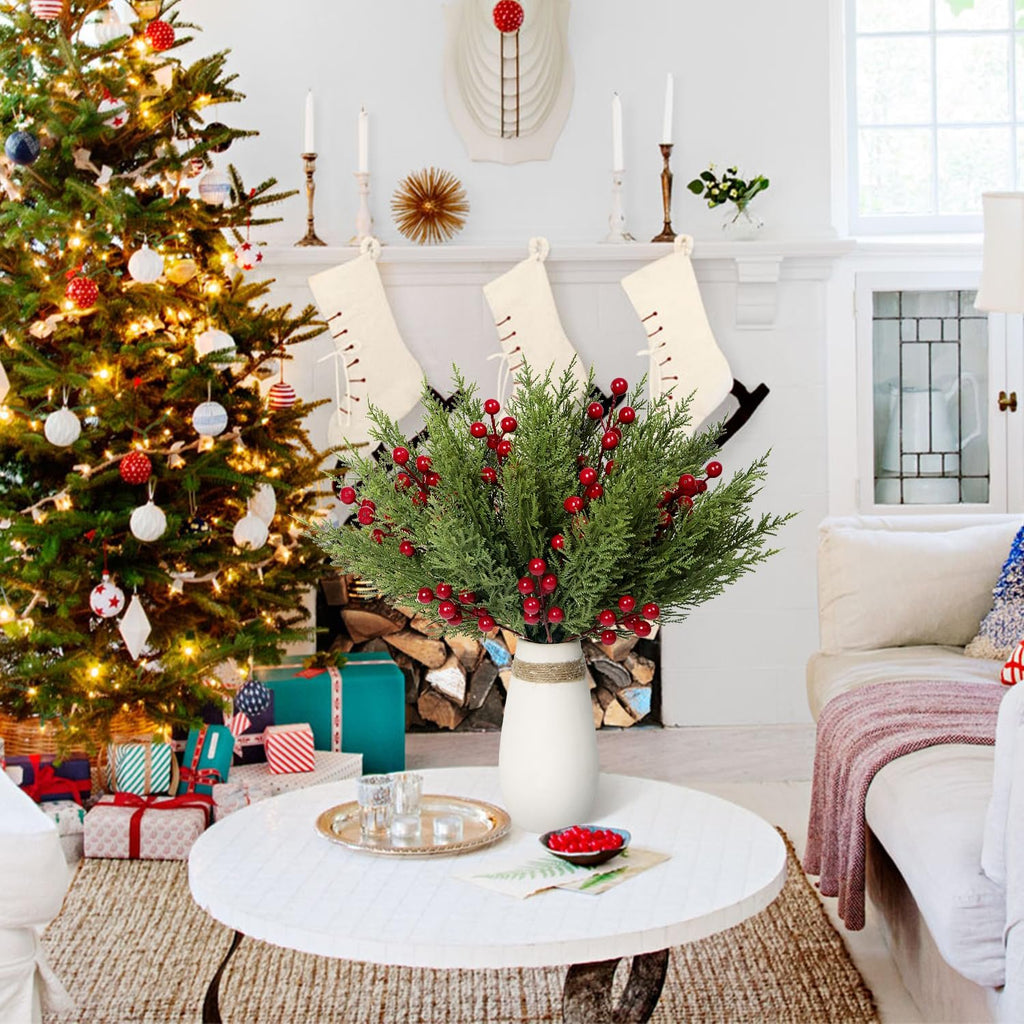 Artificial pine branches with red berries in a white vase on a coffee table, with a Christmas tree and fireplace in the background.
