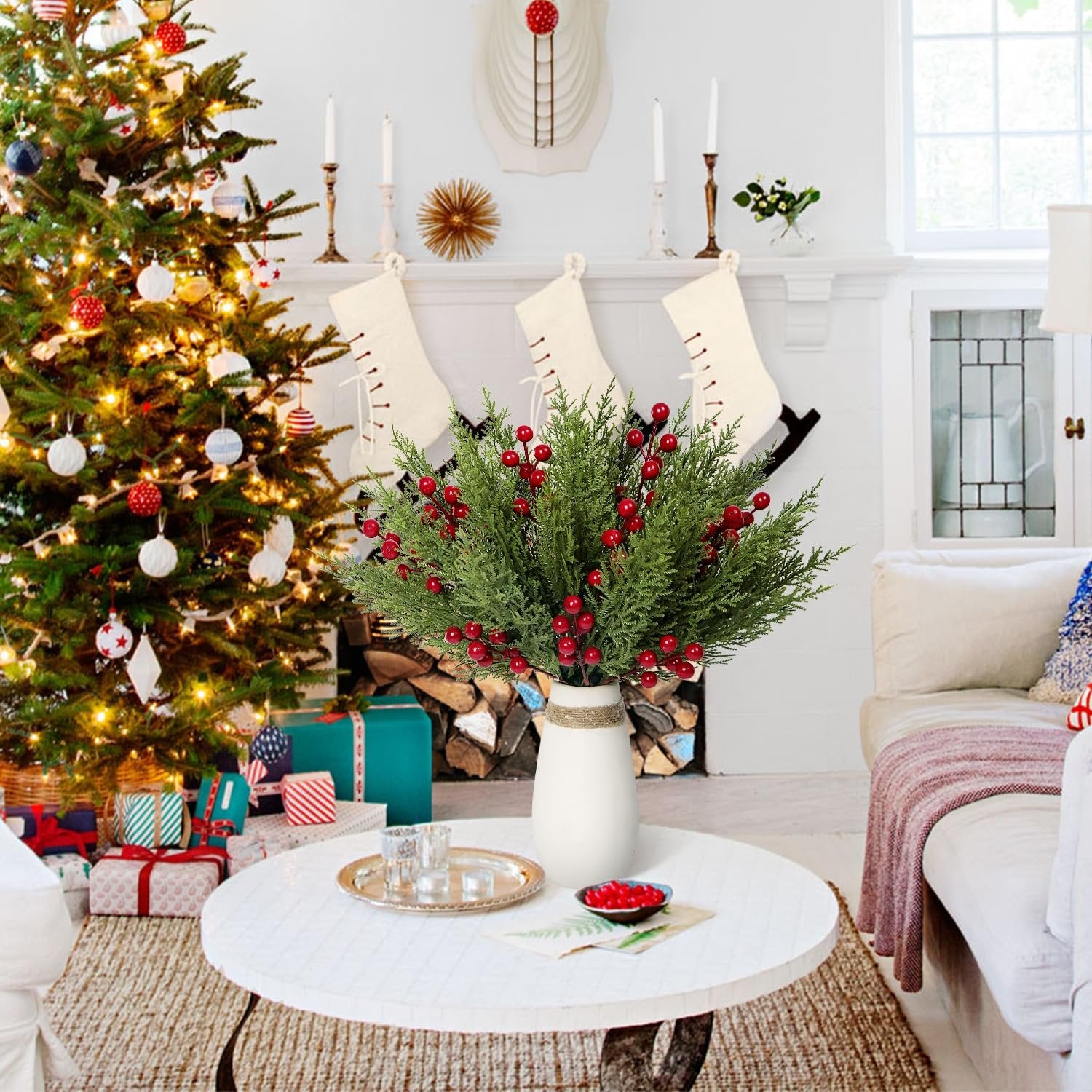 Artificial pine branches with red berries in a white vase on a coffee table, with a Christmas tree and fireplace in the background.