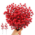 Hand holding a bunch of artificial red berry stems with two smaller stems to the left on a white background.