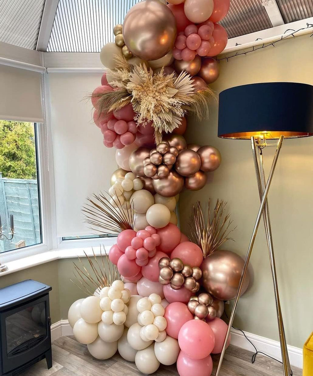 Dusty pink, rose gold, and ivory balloon garland with dried floral accents arranged along a wall next to a window and a floor lamp.