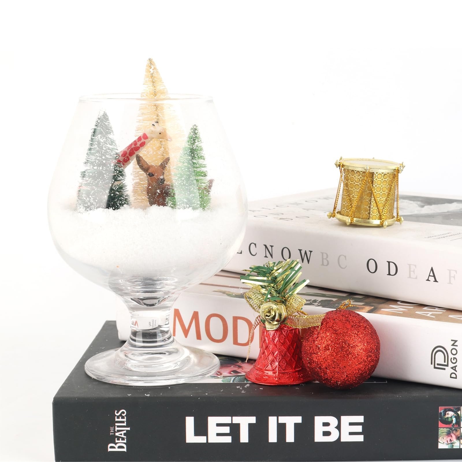 Miniature snowy Christmas scene inside a glass goblet with deer and trees, next to decorative ornaments and books.