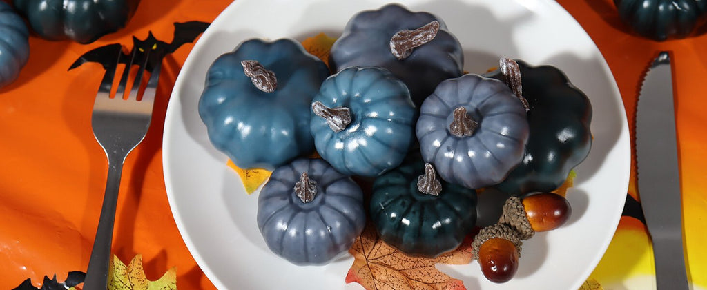 Assorted blue and gray mini pumpkins arranged on a white plate with fall leaves, fork, knife, and acorns.
