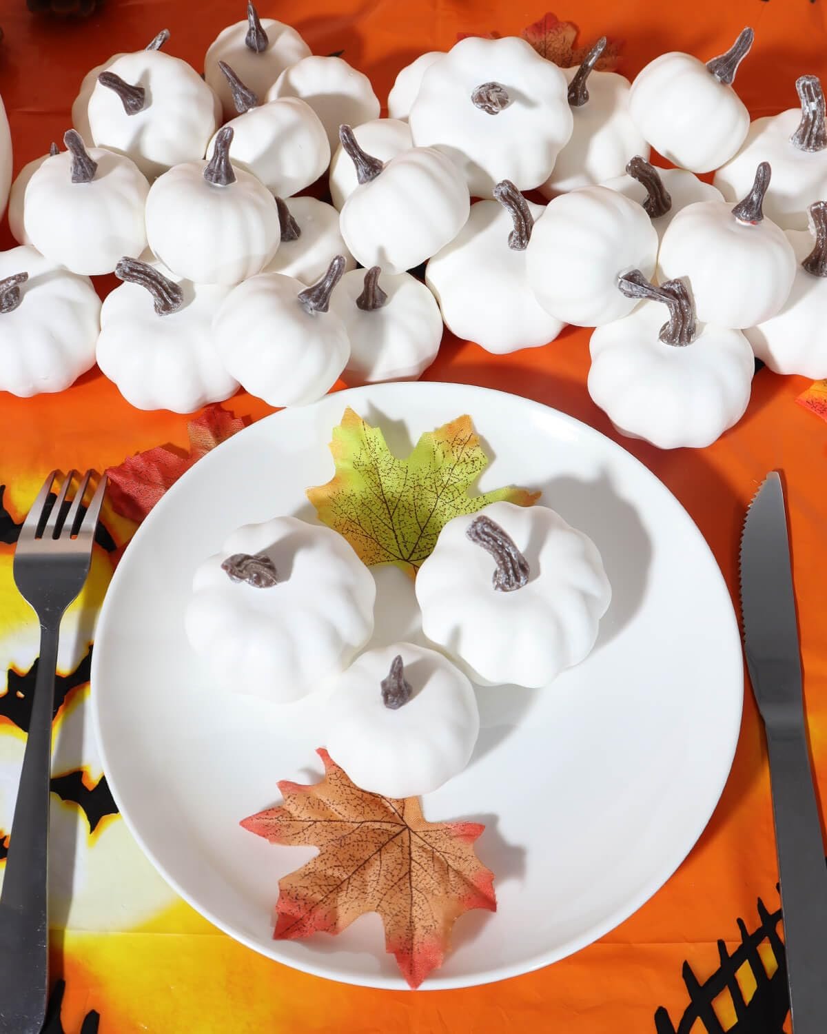 White mini pumpkins arranged on a plate with fall leaves, next to cutlery, on an orange Halloween-themed tablecloth.